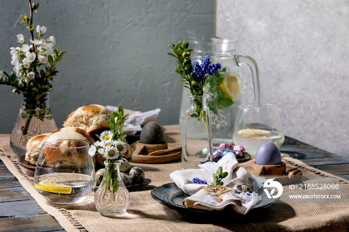 Easter table setting with colored and chocolate eggs, hot cross buns, bouquet flowers, empty ceramic plate with napkin, glass of lemonade drink on wooden table with textile tablecloth.