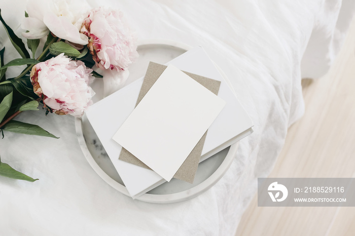 Wedding still life scene. Greeting card mockup scene, craft paper envelope, book on marble tray. Pink peony flowers on white linen table cloth. Vintage feminine styled photo, wooden floor. Flat lay.