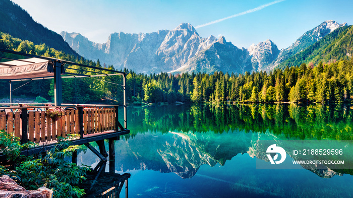 Cozy cafe on the pier. Calm morning view of Fusine lake. Colorful summer scene of Julian Alps with Mangart peak on background, Province of Udine, Italy, Europe. Beauty of nature concept background.