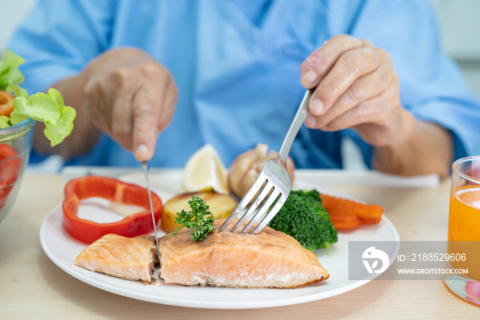 Asian senior or elderly old lady woman patient eating Salmon steak breakfast with vegetable healthy food while sitting and hungry on bed in hospital.