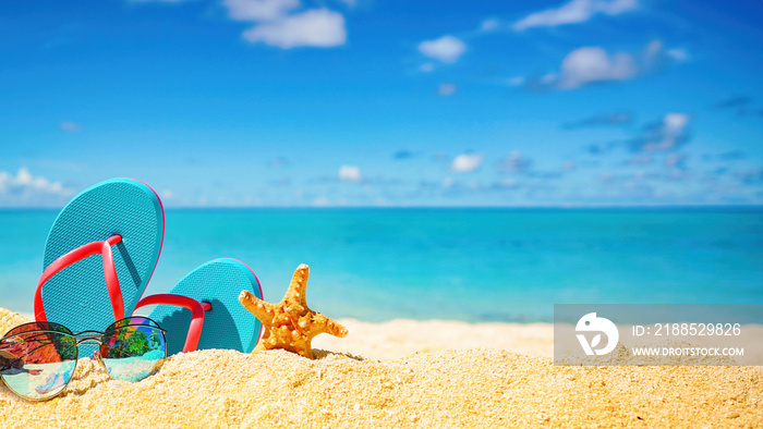 Beautiful colorful background for summer beach holiday. Sunglasses, starfish, turquoise flip-flops on sandy tropical beach against blue sky with clouds on bright sunny day.