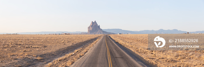 Panoramic View of a road in a dry desert with a Shiprock mountain peak in the background during a vibrant sunny sunrise.Taken at Rattlesnake, New Mexico, United States.