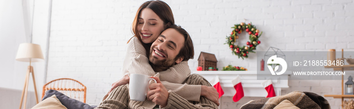 joyful couple embracing with closed eyes in living room with blurred christmas decoration, banner