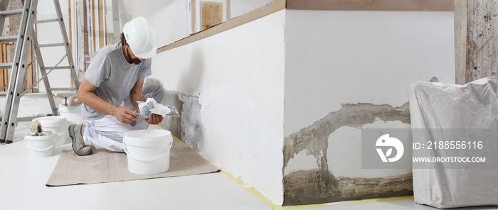 plasterer man at work, take the mortar from the bucket with trowel to plastering the wall of interior construction house site and wear helmet, panoramic image