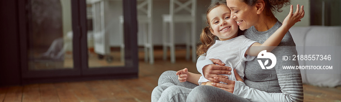 Jolly mother is hugging with little girl while doing workout together in cozy living room