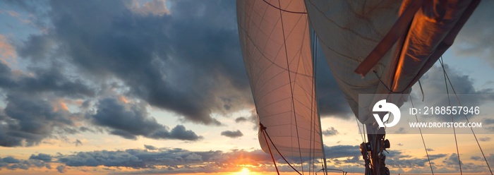 Yacht sailing in an open sea at sunset. Close-up view of the deck, mast and sails. Clear sky after the rain, dramatic glowing clouds, golden sunlight, waves and water splashes, cyclone. Epic seascape