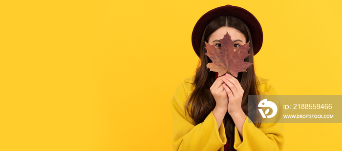 funny teen girl in hat and coat hiding behind autumn maple leaf on yellow background, season. Autumn fall child for poster design. Banner header, copy space.