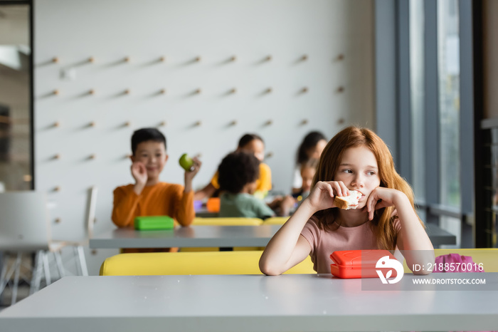 sad redhead girl eating sandwich near schoolkids on blurred background