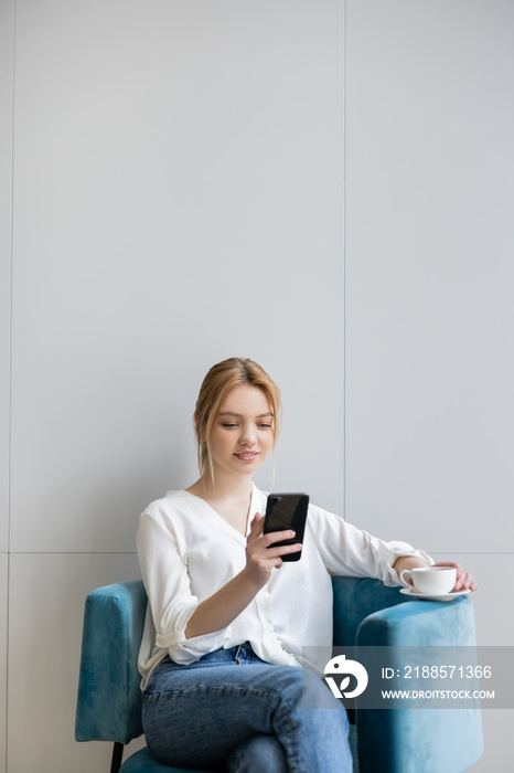 Young woman using smartphone and holding coffee on armchair at home.