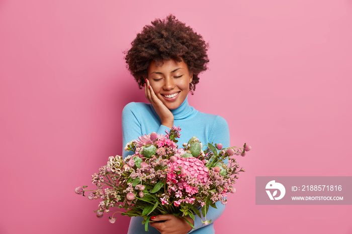 Photo of pleased touched young Afro American woman got gift for anniversary, carries big bouquet of 