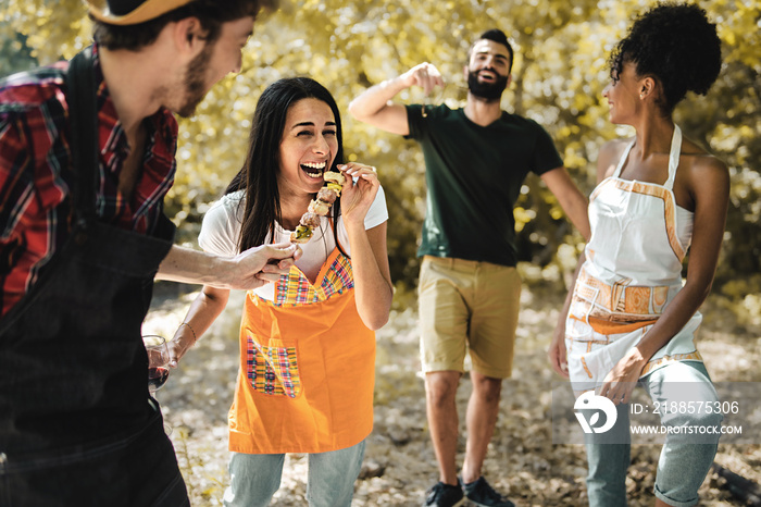 Multiethnic group of young friends carefree at picnic joking together and eating meat skewers grille