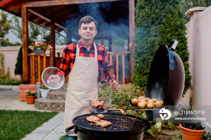 Man in a shirt and an apron is cooking barbecue meat on a grill in his home garden.