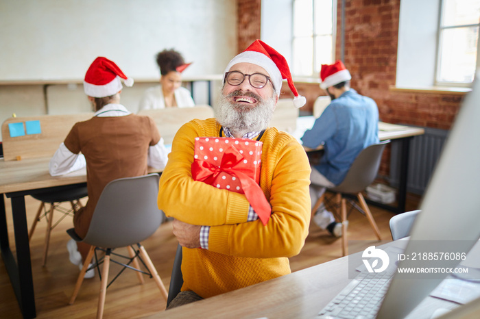 Ecstatic senior businessman in Santa cap embracing giftbox with xmas present while sitting by workpl