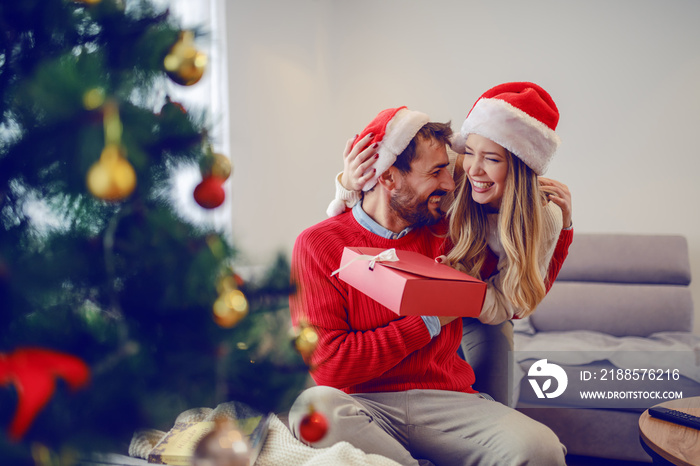 Overjoyed handsome bearded man sitting on sofa in living room, holding gift and hugging with his gir