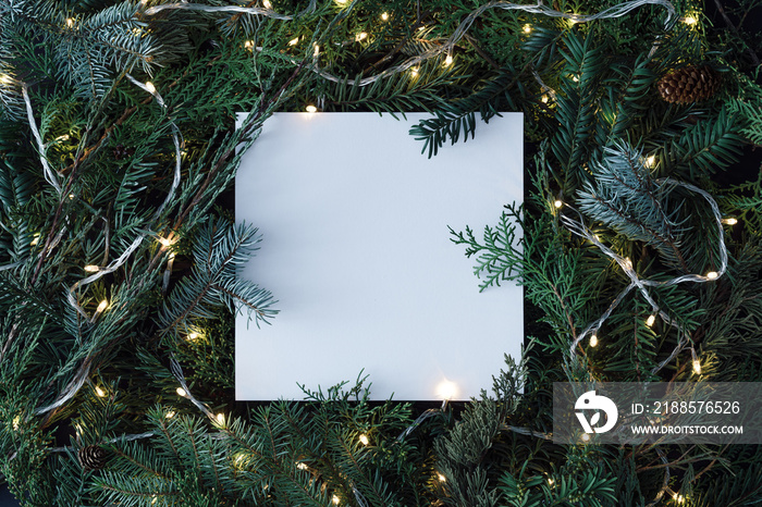 Creative layout made of Christmas tree branches with paper card note and christmas lights. Flat lay.