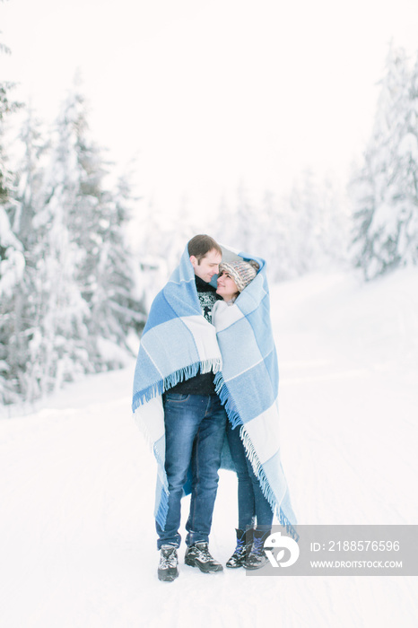 Happy lovely couple walking in winter frosty forest and covering with blue chekered wool blanket