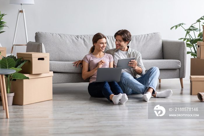 Cheerful european millennial guy in glasses and female sit on floor with laptop and tablet in room i