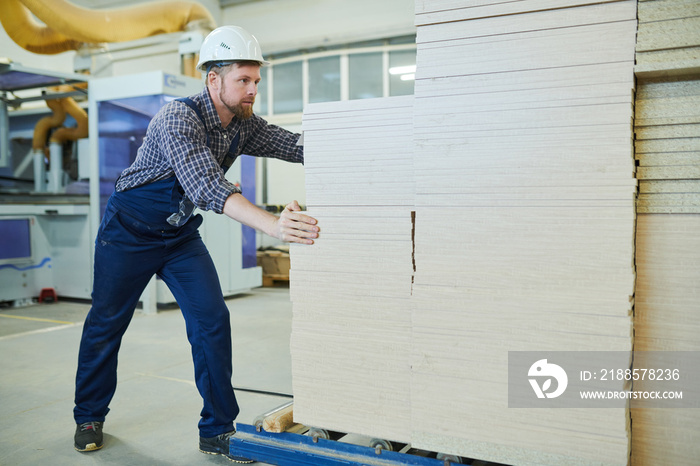 Serious bearded strong worker in hardhat and overall pushing stack of wooden pieces on roller convey