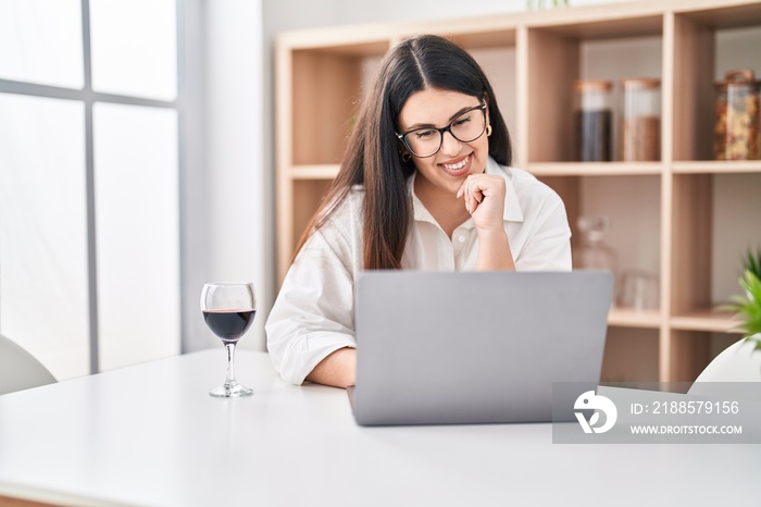 Young hispanic woman using laptop drinking wine at home