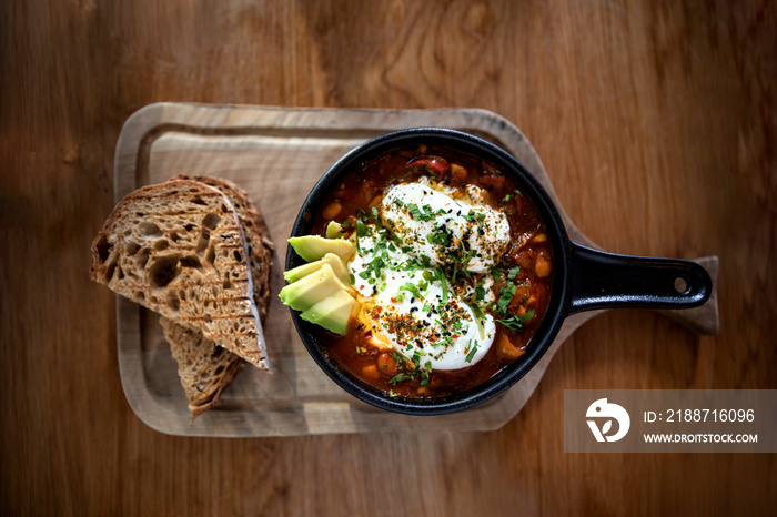 Top view. Shakshuka with two poached eggs and avocado in a black pan and bread aside on a wooden board. Middle eastern traditional dish. Flat lay