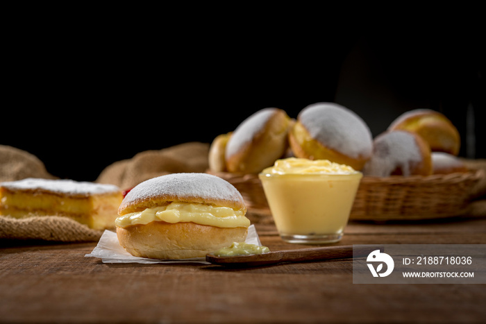 cream filled donut with suggar icing on wood with other donuts and a cream Bowl on background