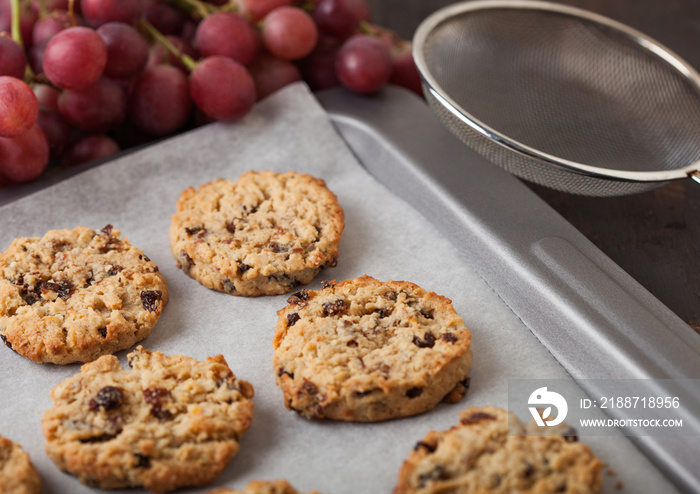 Homemade organic oatmeal cookies with raisins in baking tray with grapes and backing mesh on wooden background. Top view