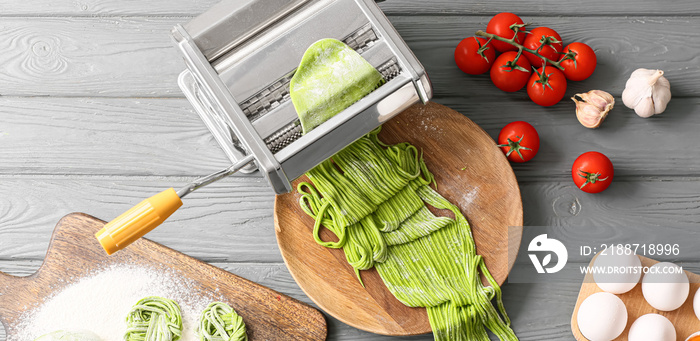 Pasta machine with green dough and ingredients on wooden background, top view