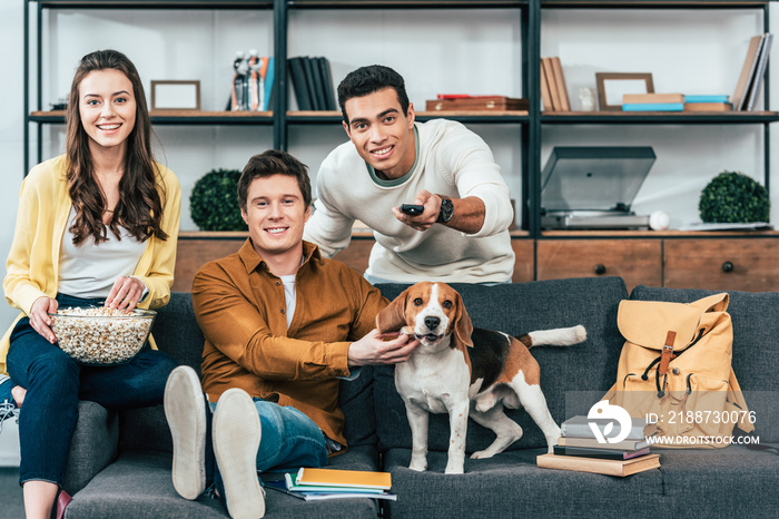 Three smiling multicultural friends with dog and popcorn sitting on sofa and watching tv