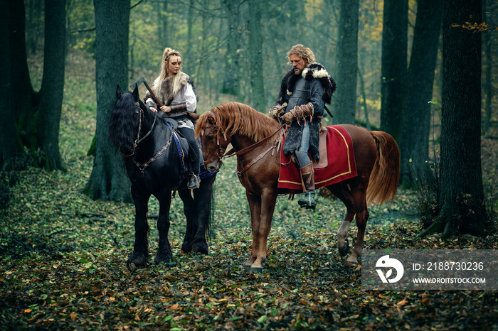 Warriors Woman an Man on horses in the woods. Scandinavian vikings riding horse with axes in hands. Traditional clothes with fur collar. Reconstruction of a medieval scene