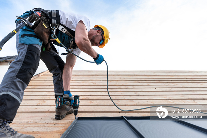 A roofer with a safety harness and tool belt is working with a electric screwdriver on the roof. He is anchoring the metal roofing with a screws.