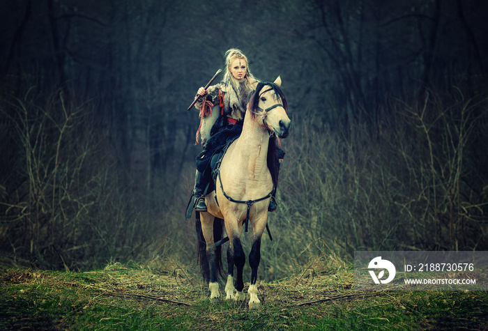 Warrior Woman on a horse in the woods. beautiful viking riding horse with ax in hand, blonde hair in a traditional clothes with fur collar, war, forest. Reconstruction of a medieval scene