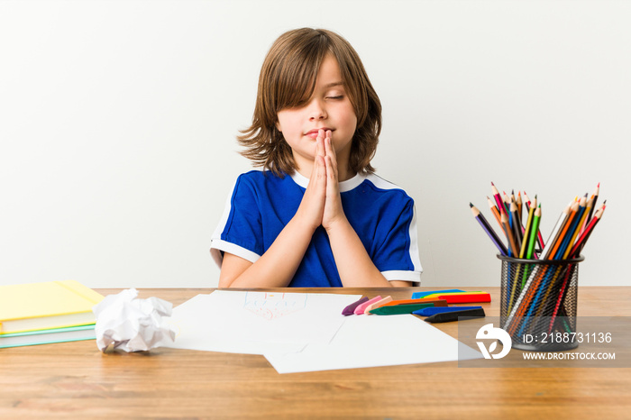 Little boy painting and doing homeworks on his desk holding hands in pray near mouth, feels confident.