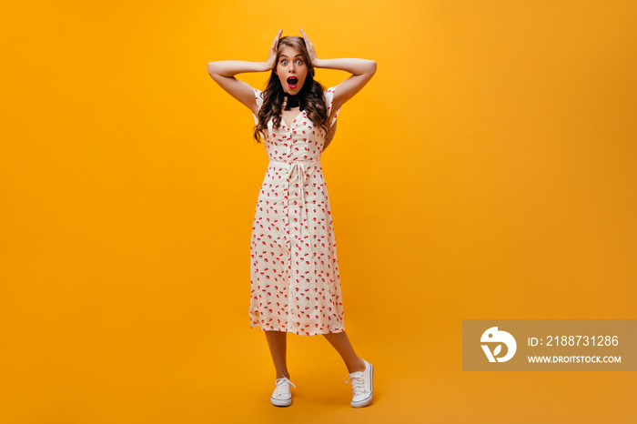 Shocked woman in midi dress looks into camera. Curly girl in white summer outfit and sneakers posing on orange background..