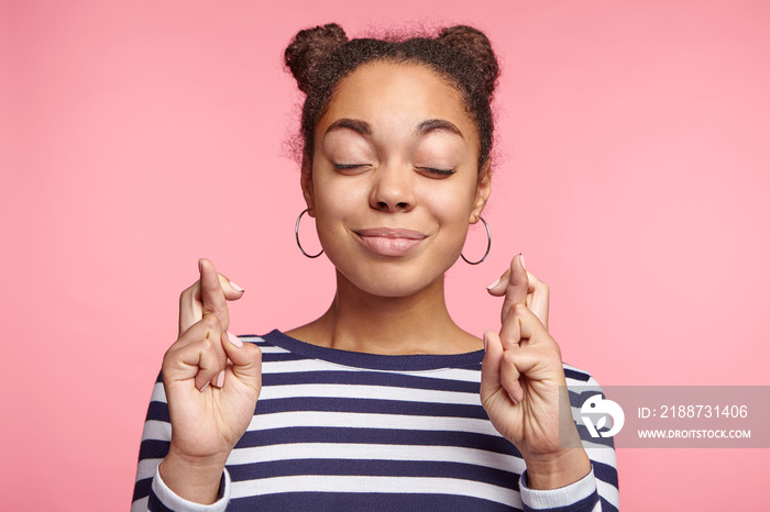 Isolated shot of attractive coquette female model with dark skin closes eyes and crosses fingers in anticipation, believes for good luck, isolated over pink studio background. Woman gestures indoors