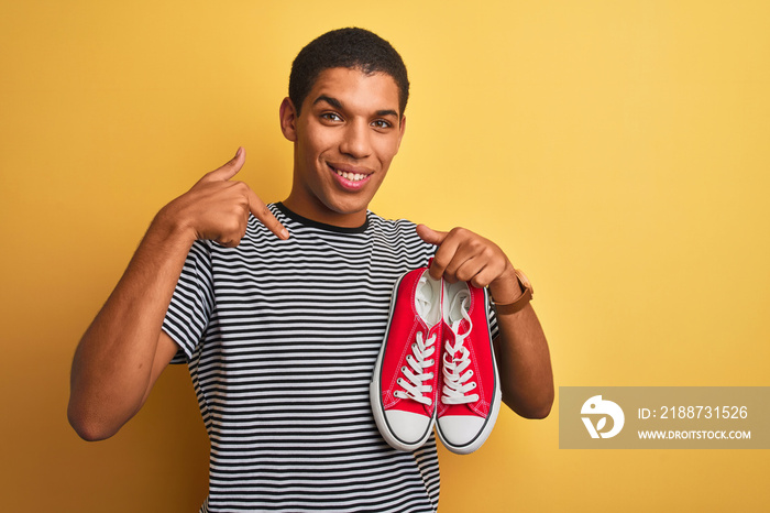 Young handsome arab man holding casual sneakers standing over isolated yellow background with surprise face pointing finger to himself