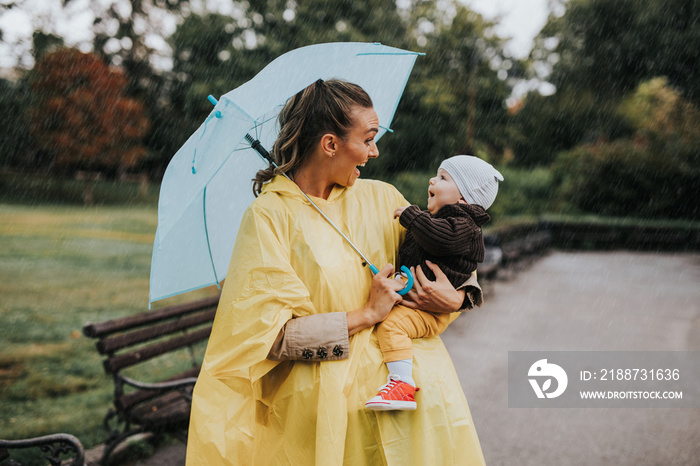 Beautiful and happy middle age woman with her baby walking in city park on rainy day..