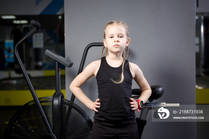 Small attractive caucasian child using exercise bike in the gym. Fitness. A little athlete using an air bike for a cardio workout at the crossfit gym. Athletic girl posing near the cycling machine