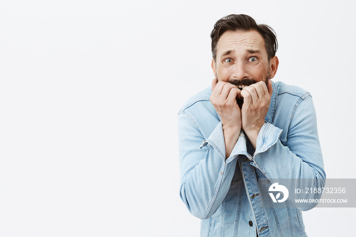 Gosh not again, most scary fear come alive. Portrait of shook and terrifying anxious guy, biting fingernails and stooping while standing over gray background in denim jacket, being frightened to death