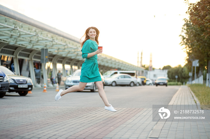 Young happy woman runs across the crosswalk. Hot coffee in a plastic glass. Green dress. The concept of business, lifestyle and technology.