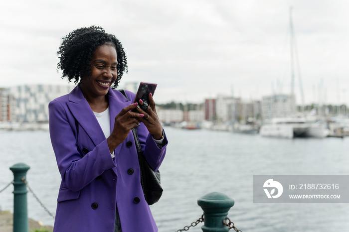 Smiling woman using smart phone by river in city