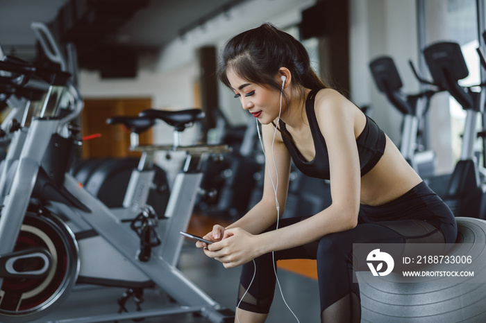 Young asian woman athlete listening music with smartphone relax after break workout and cardio sport exercise at fitness gym on yoga ball for healthy lifestyle