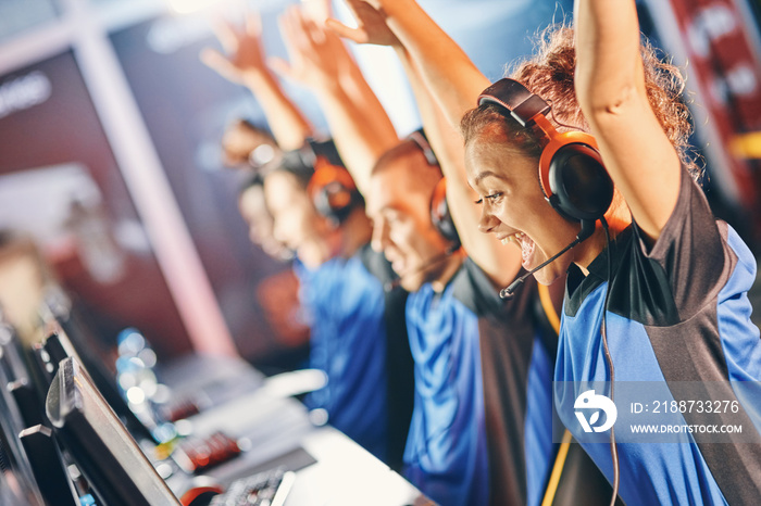 Side view of a excited mixed race girl, female cybersport gamer raising hands up, celebrating success with her team, participating in esports tournament