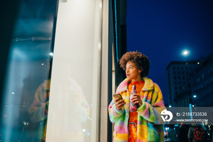 Young mixed race woman using reusable water bottle and smartphone walking outdoor at night