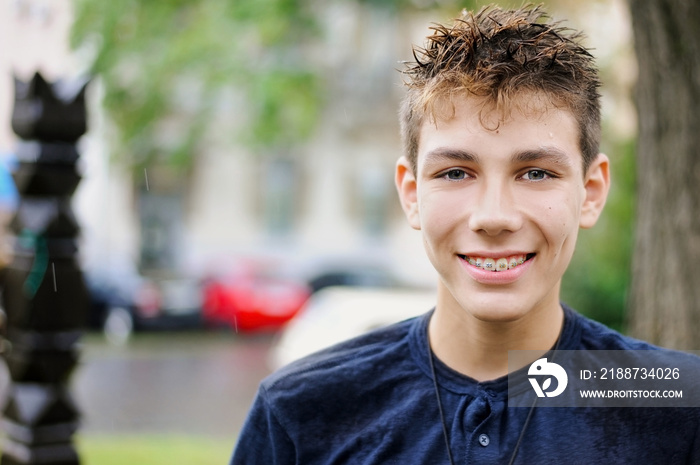 A nice boy walking in the rain on the street. He has a wet hair but he is still happy and smiles. Braces on his teeth are clearly visible