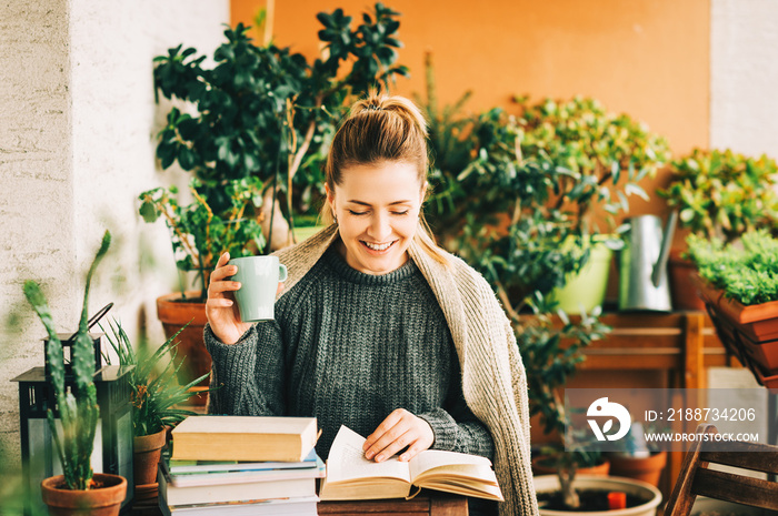 Young beautiful woman relaxing on cozy balcony, reading a book, wearing warm knitted pullover, holding cup of tea or coffee