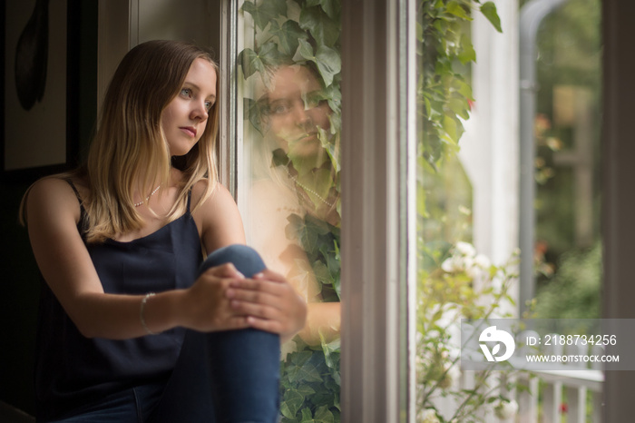 Junge Frau sitzt verträumt am Fenster - Blondes Mädchen sitzt auf der Fensterbank und schaut verträumt hinaus