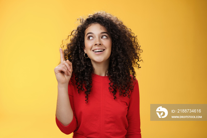 I got idea. Excited happy attractive woman with curly hair in red dress raising index finger in eureka gesture smiling amused, looking at upper left corner, sharing plan or suggestion over yellow wall