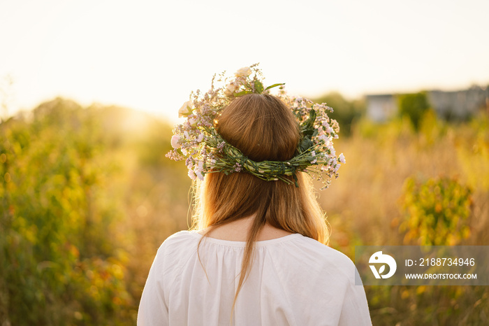 Summer lifestyle portrait of beautiful young woman in a wreath of wild flowers. Standing back in the flower field, hands to the side. Romantic mood. Nature lover