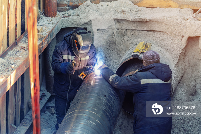 Welder welding water or gas steel pipeline with assistant worker in trench. City underground utilitites renewal and replacemen