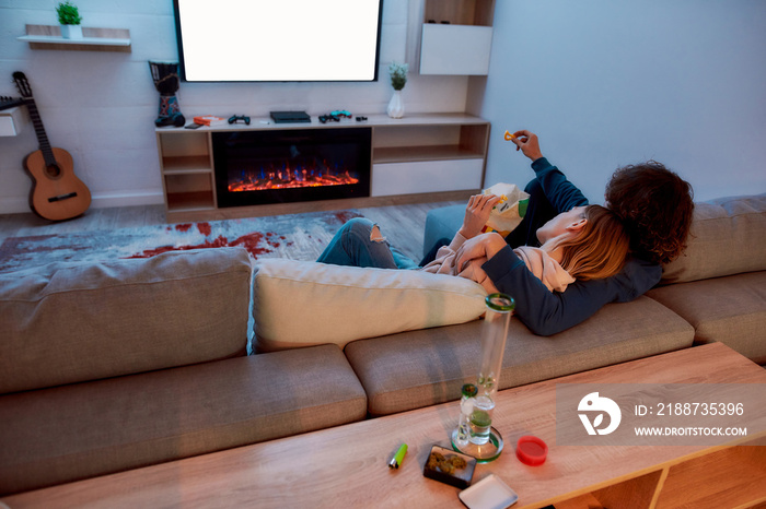 Back view of young couple watching TV together, eating snacks after smoking marijuana from a bong or glass water pipe. They relaxing on the couch in the evening at home